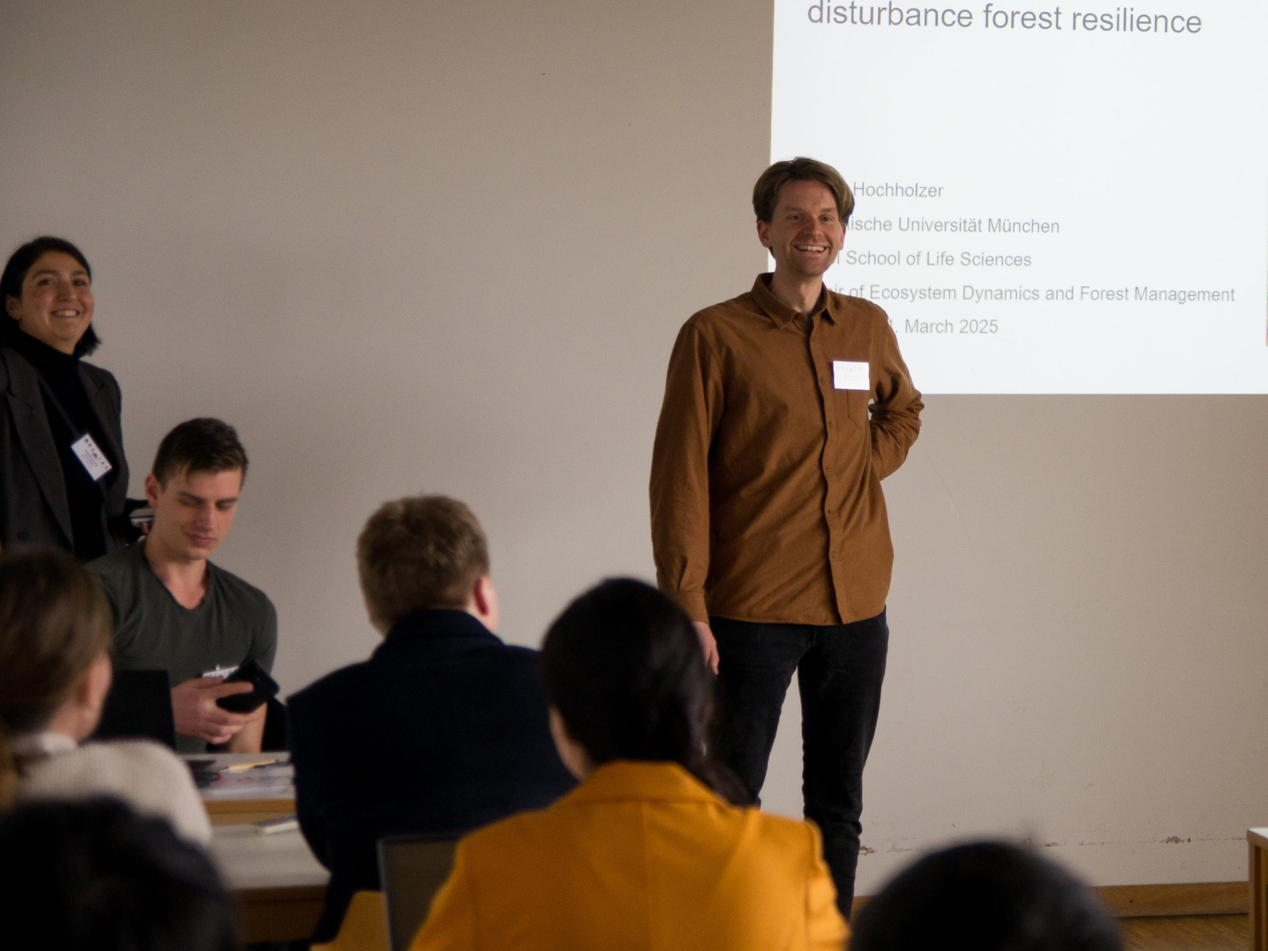 A man in a brown button-up smiles in front of a projection, partially covering the text behind him. Other people nearby are also smiling. The part of the screen that can be read says: "disturbance forest resilience. Hochholzer, Technische Universität München, School of Life Sciences, Chair of Ecosystem Dynamics and Forest Management, March 2025".