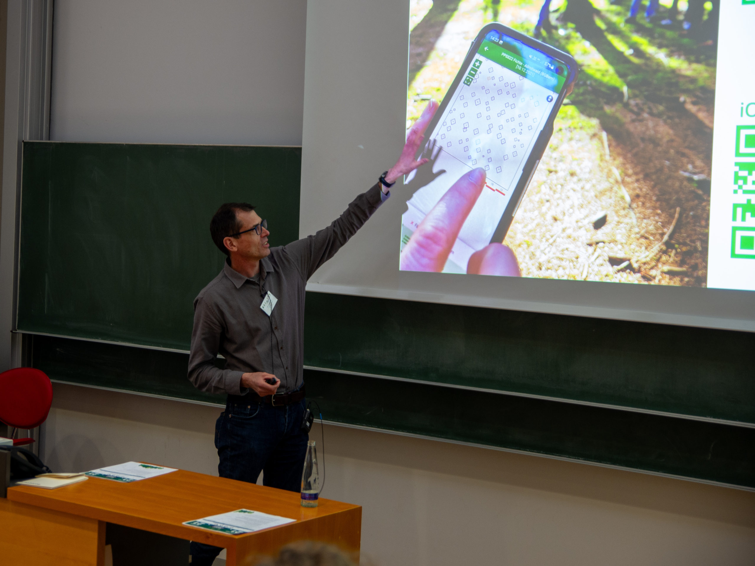 A dark-haired man in glasses and a grey button-up gestures at a projector screen. On the screen is a mobile device, showing an app with zoom-buttons and various diamond-shapes on a white background. A green bar along the top reads "PPSS22 Fichte - Alimitwald (BGBern) [10.12.2021]"