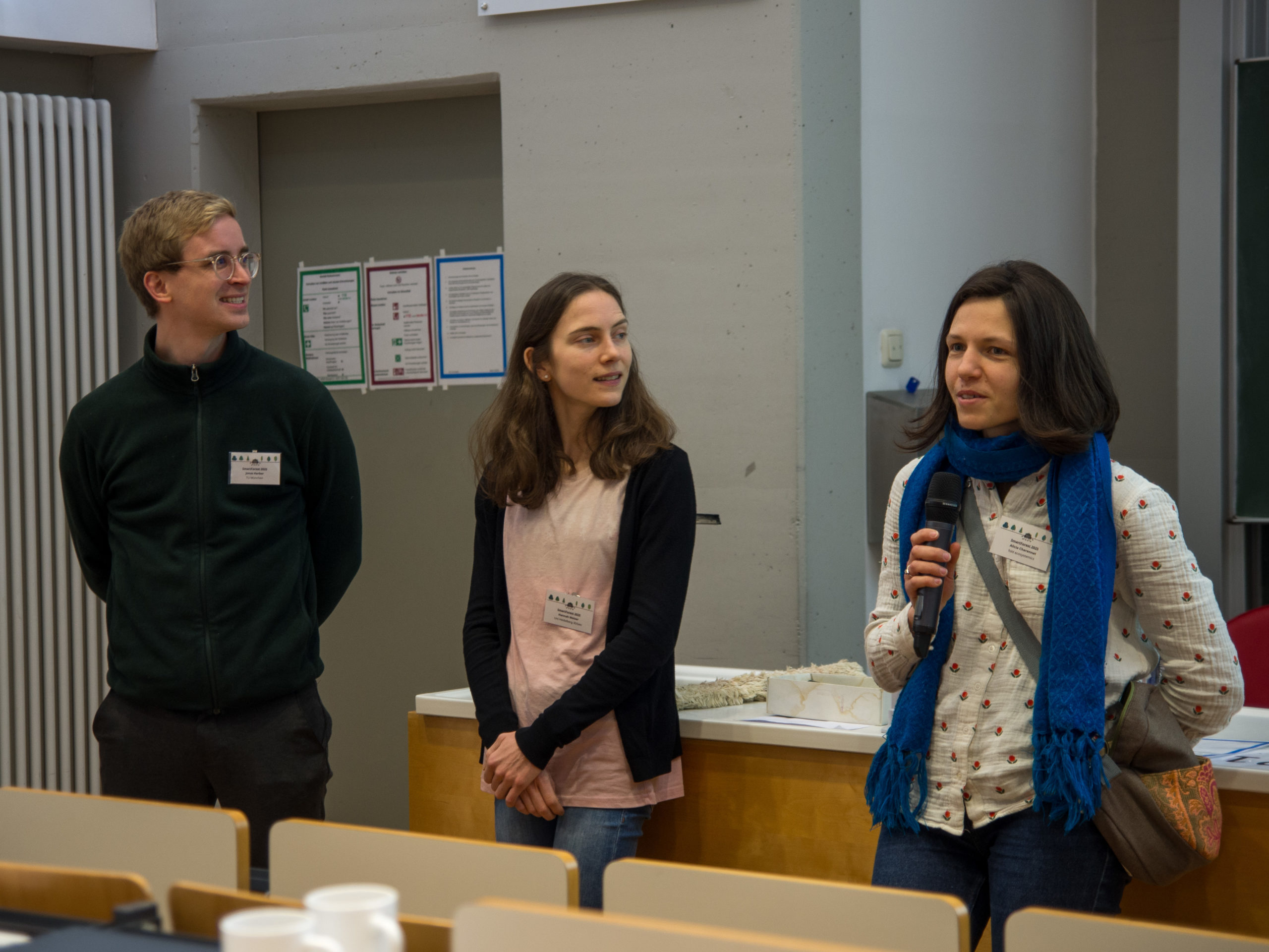 Three people stand in a lecture hall, facing the room. To the left, Jonas Kerber (a blond man in glasses and a dark zip-up sweatshirt) and Hannah Weiser (a brunette sporting a pink top with a dark vest) look over to the right, where Alicia Charennat (a dark-haired woman in a light-coloured blouse with flower motif and a blue scarf) speaks into the microphone.