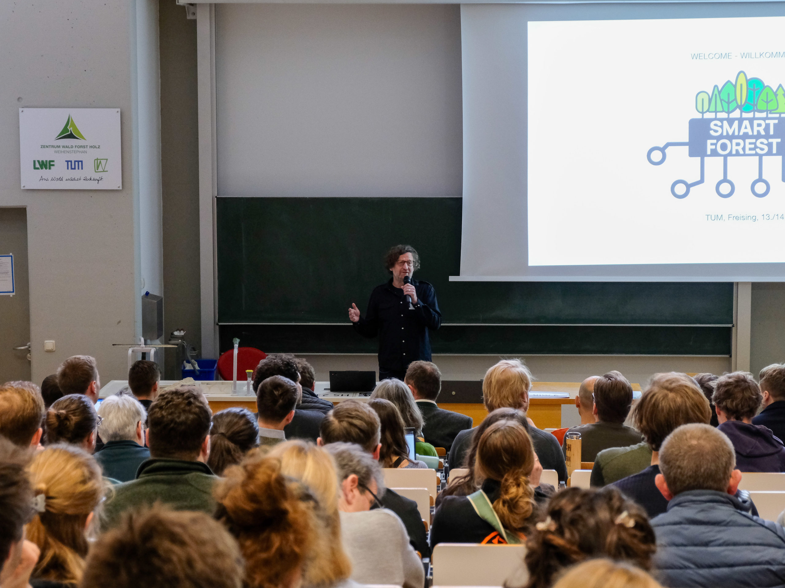 Dr. Werner Rammer addresses a full lecture hall. To the left is a sign of the "Zentrum Wald Forst Holz Weihenstephan", while on the right is a projected slide welcoming people to SmartForest.