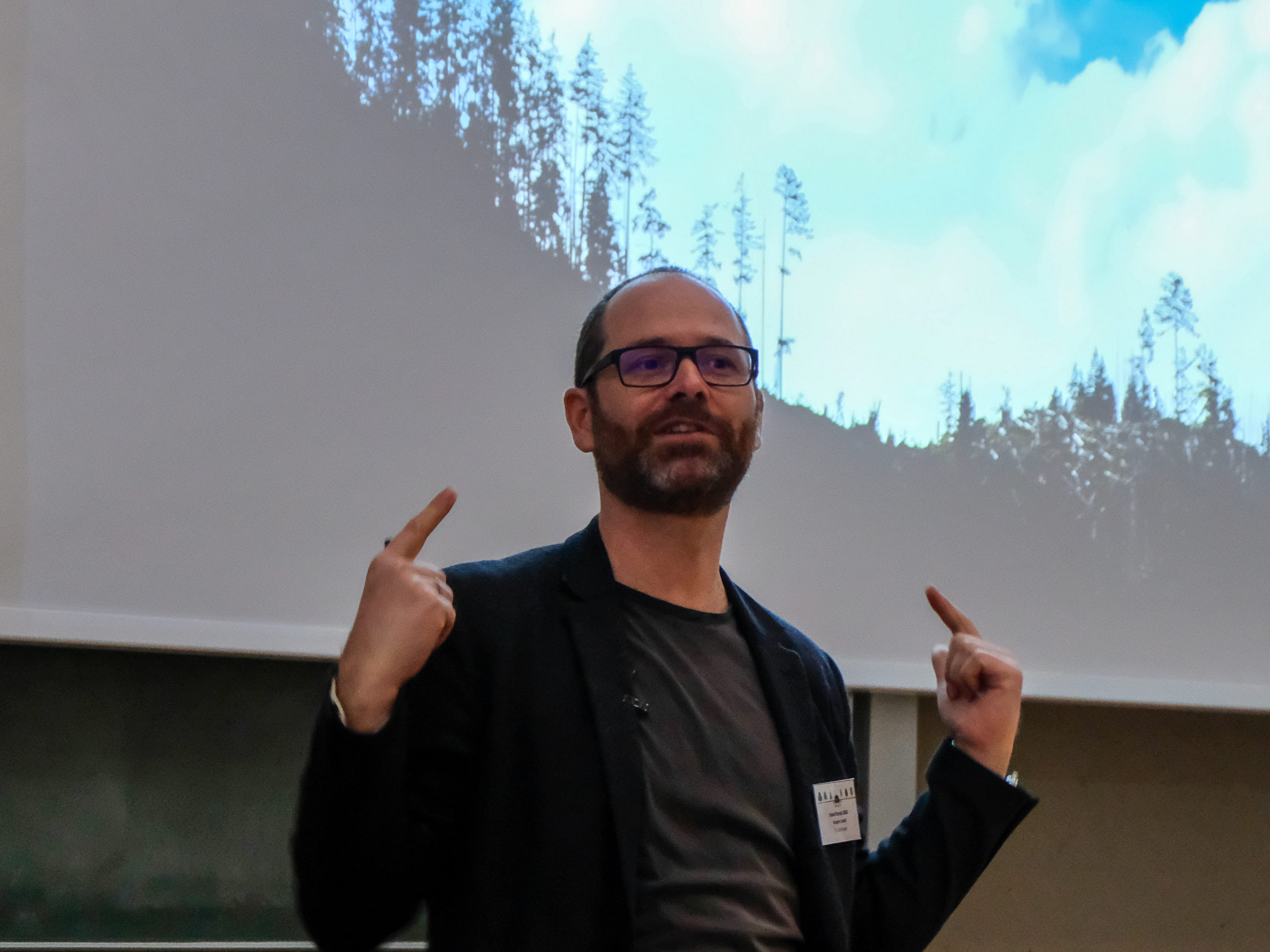 Prof. Dr. Rupert Seidl, a bespectacled man with short dark hair and a beard, points up towards his head with both fingers as he addresses the crowd. Behind him is a projection of a forest.
