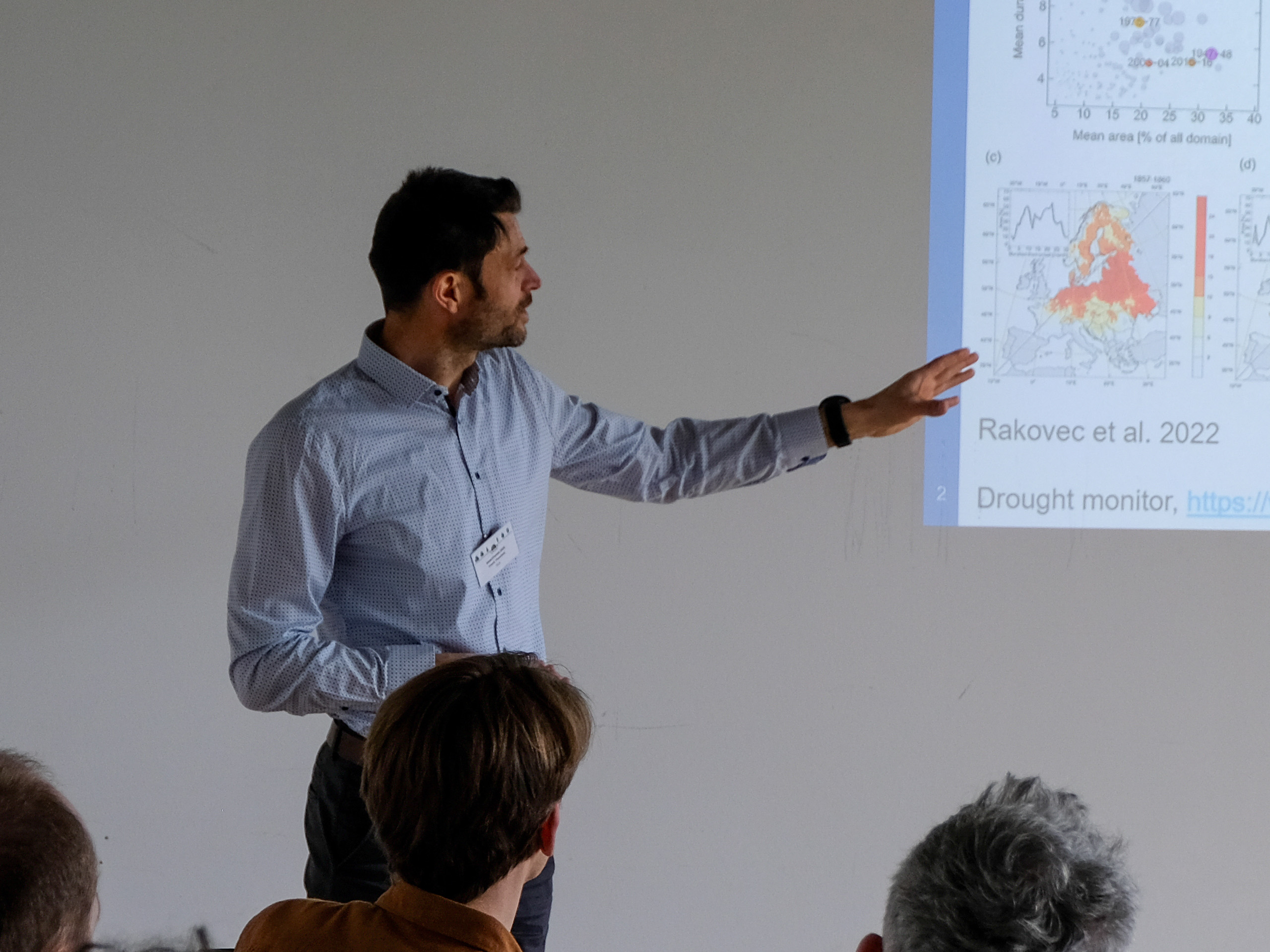Dr. Frank Thonfeld, a dark-haired man with a stubbly beard and light blue button-up shirt, gestures at a projected slide showing location data. Only part of the slide is visible, including the text "Rakovec et al. 2022" and "Drought monitor".