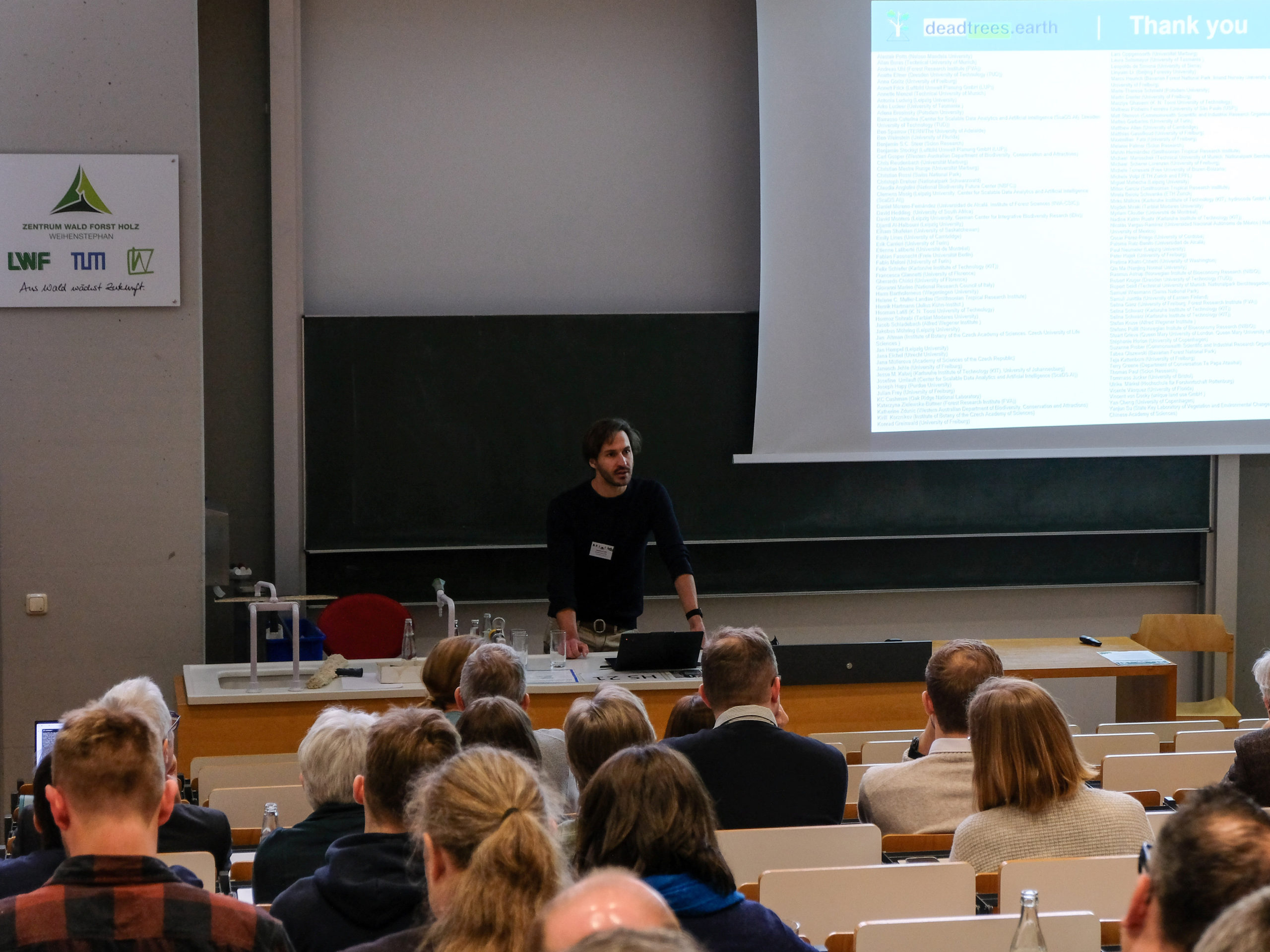 A crowd of people watches a presentation on deadtrees.earth in the main lecture hall during SmartForest 2025.