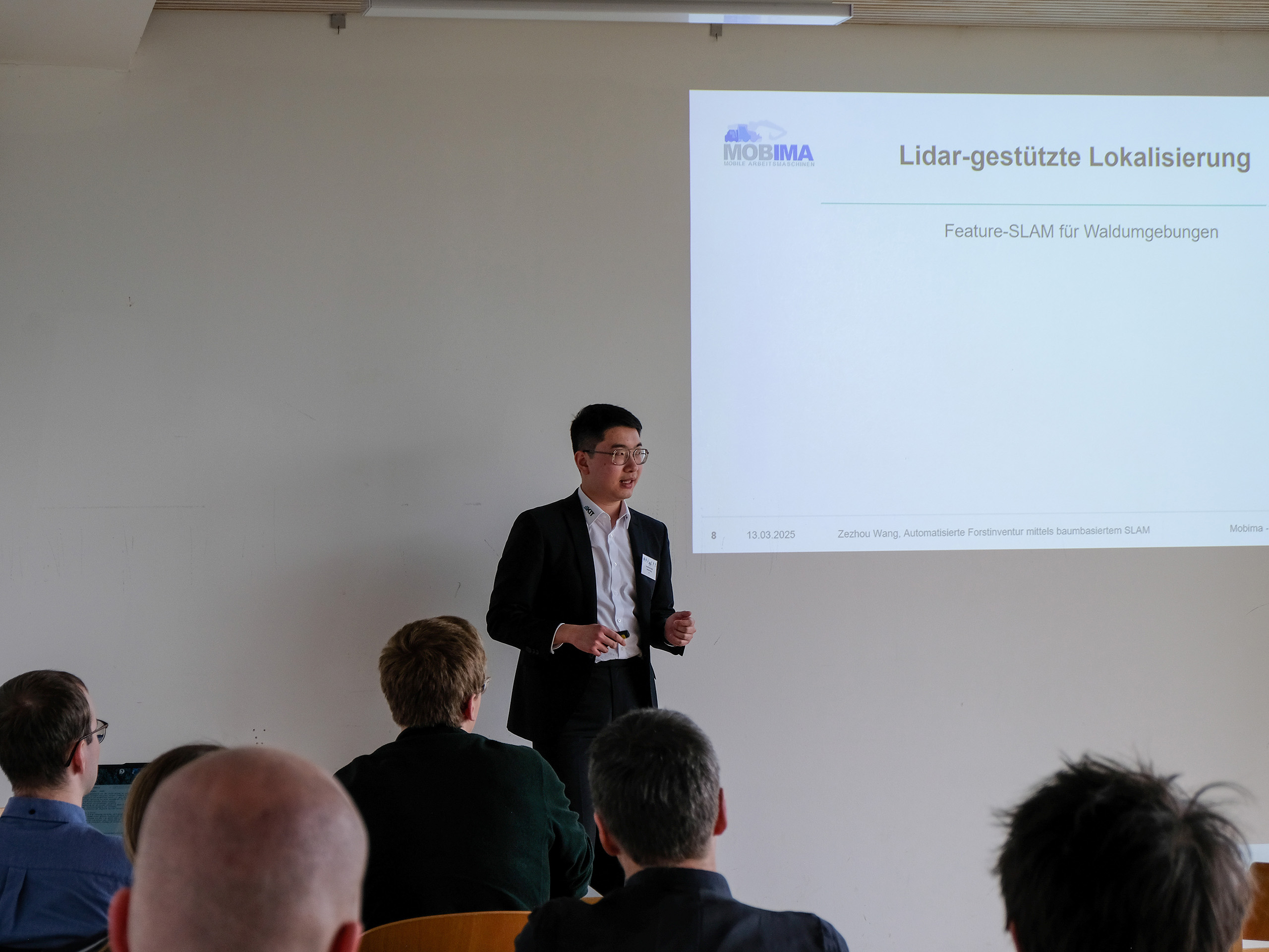Zezhou Wang, a dark-haired Asian man in a smart suit and glasses, presents a MOBIMA (Mobile Arbeitschmaschinen) slide titled "Lidar-gestützte Lokalisierung. Feature-SLAM für Waldumgebungen" to a crowd of interested professionals at SmartForest 2025.