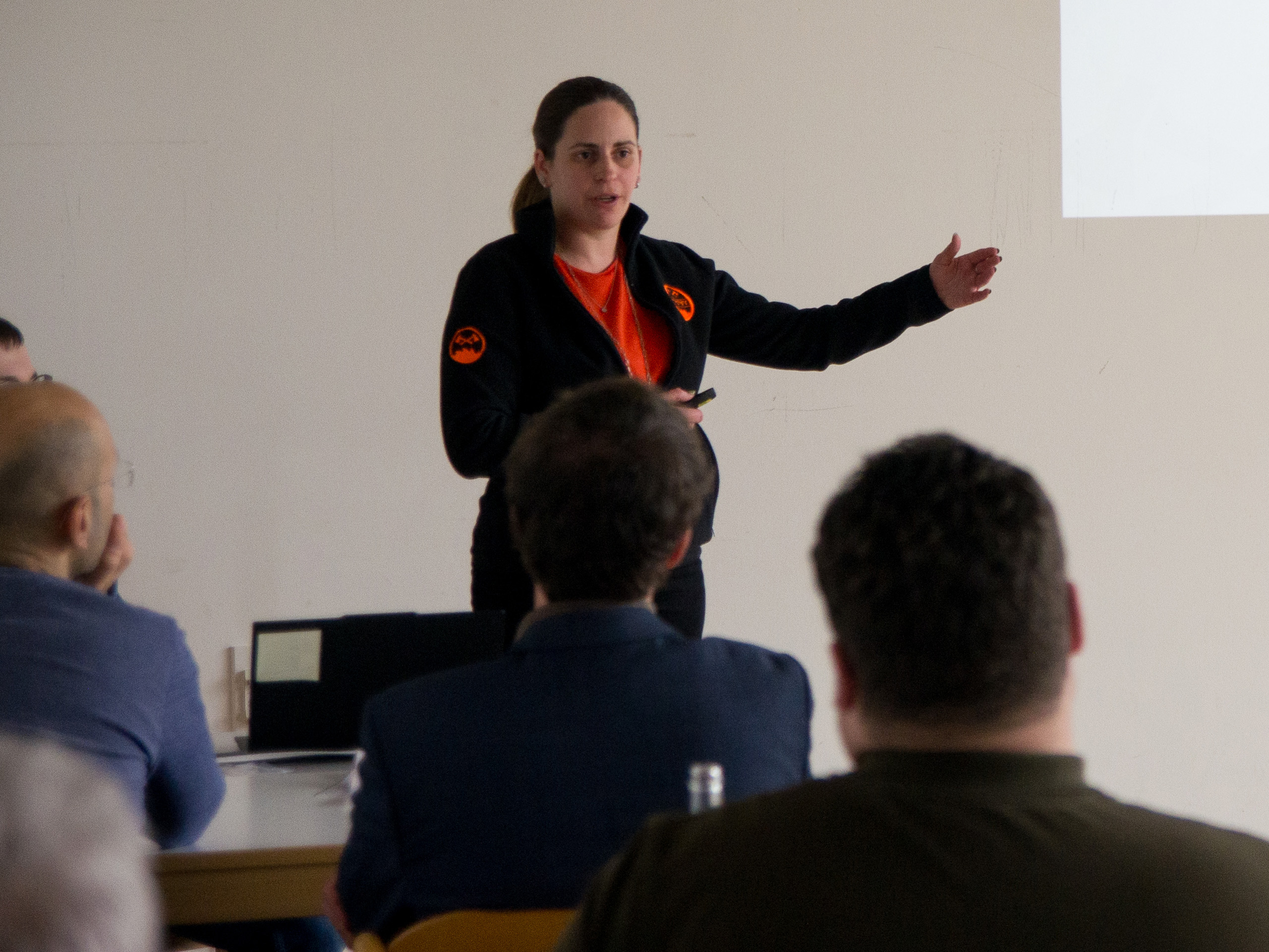 A woman with a ponytail, sporting a black-and-orange outfit featuring the Forestify logo (two crossed axes over the top half of a circular sawblade) lectures to a crowd in a classroom.