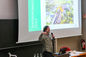 Dr. Werner Rammer of the TU Munich welcomes the crowd to SmartForest 2026. There is a large projection screen behind him, on which u0022ZENTRUM WALD FORST HOLZu0022 and the wetransform logo re visible, alongside an aerial picture of the conference area, with an arrow indicating the building.