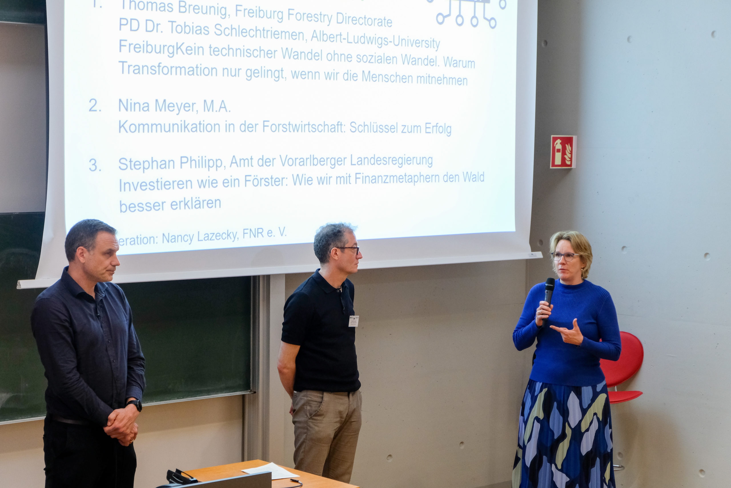 Thomas Breunig, Nina Meyer, and Stephan Philipp exchange ideas on communication in front of a crowd at SmartForest 2026. In the background, their respective presentations are listed on a projected slide.