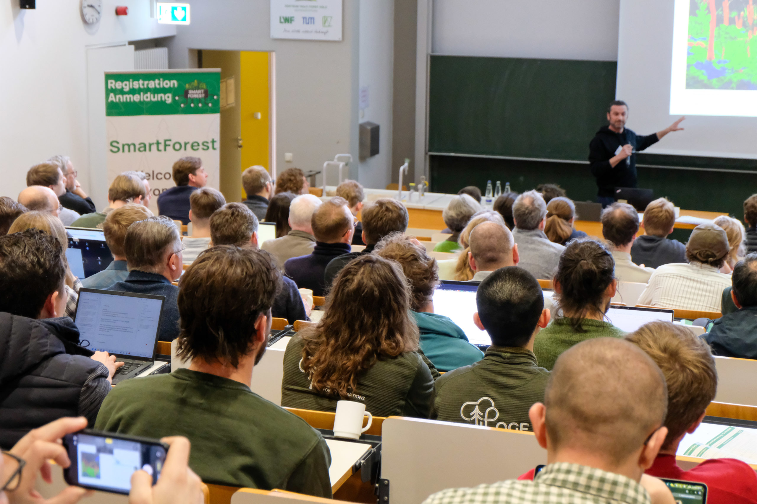 A crowd dressed primarily in green, seen from the back, seated in a full lecturing hall during a presentation at SmartForest 2026. Several people have their laptops open in front of them and one person is taking a picture with a  smartphone.