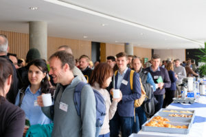 A crowd of attendees fills the foyer at SmartForest 2026, chatting over coffee. To the right is a long table bearing coffee urns, cartons of milk, crates of croissants and other baked goods.