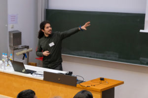 Simone Massaro of Q-ForestLab at Ghent University stands in front of a blackboard, gesturing as he addresses the seated crowd in the auditorium at SmartForest 2026