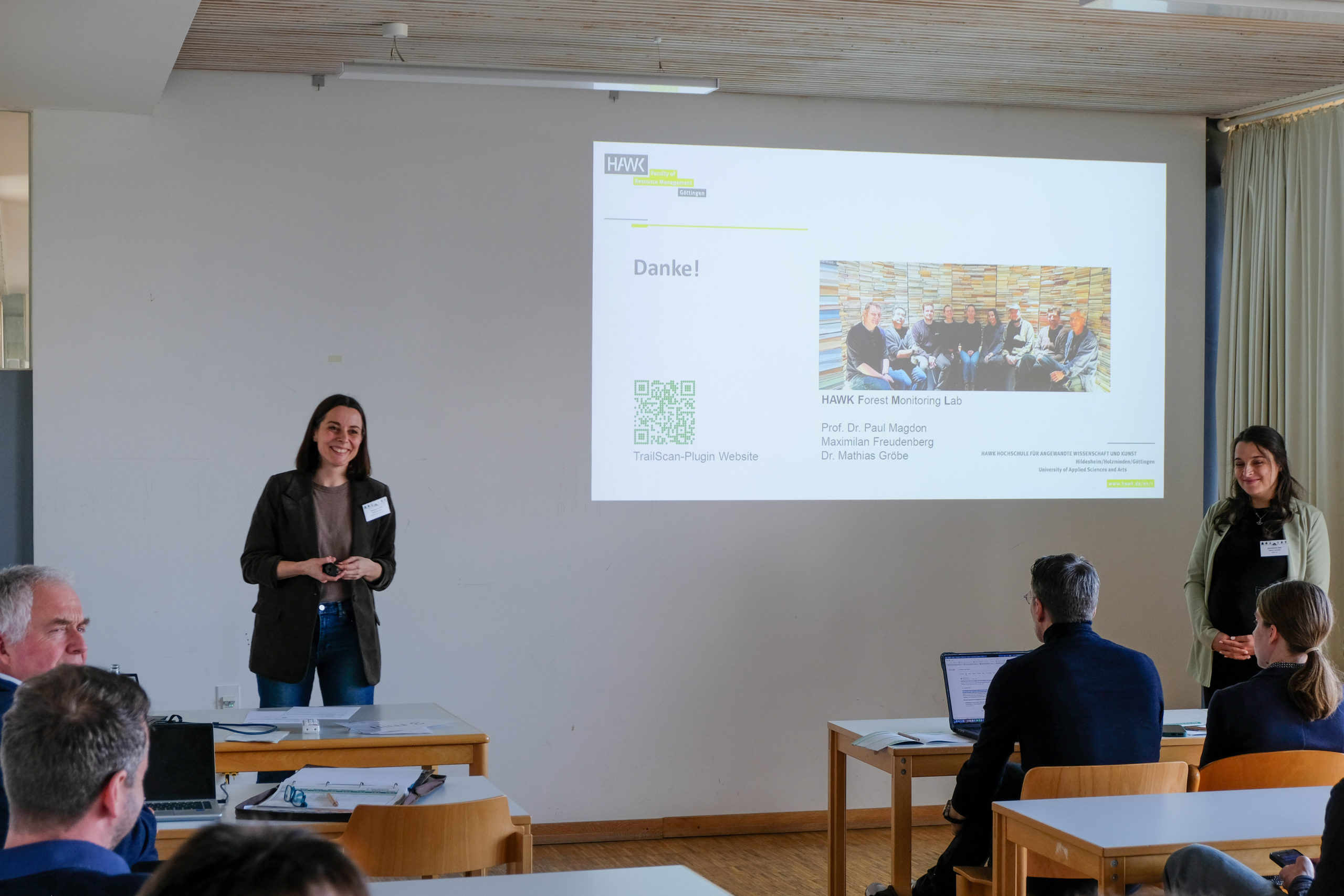 Tanja Kempen of HAWK smiles at the crowd gathered for her presentation “TrailScan: QGIS-Plugin zur Kartierung von Rückegassen in Laserscanning Daten”, the final slide of which is projected on the wall. To the right of the projection stands session moderator Nancy Lazecky.