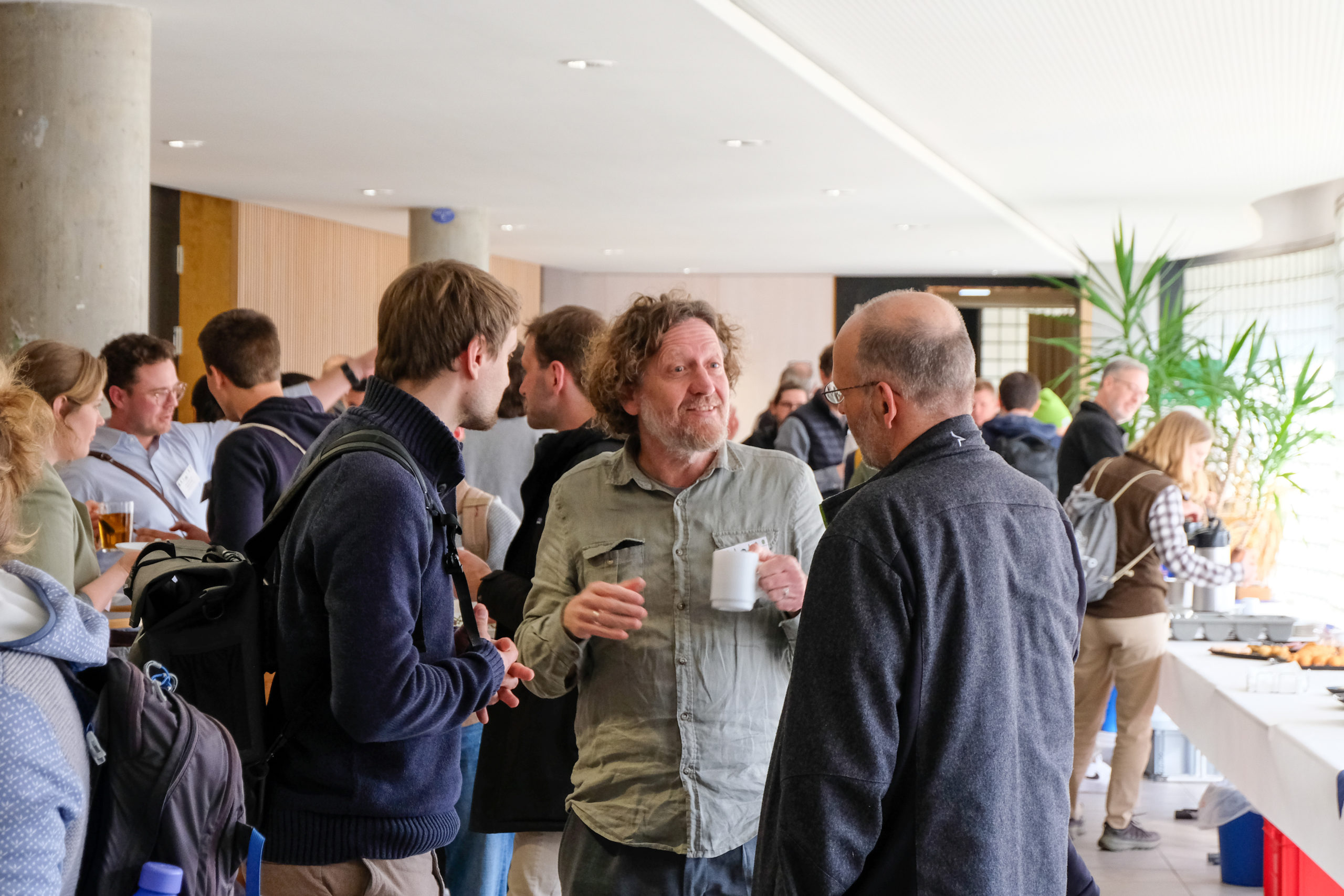 Dr. Werner Rammer of the TU Munich talks to other attendees in the pleasantly crowded foyer during SmartForest 2026. Many people have cups of tea or coffee in hand and people are picking out pastries from a long table off to the right.