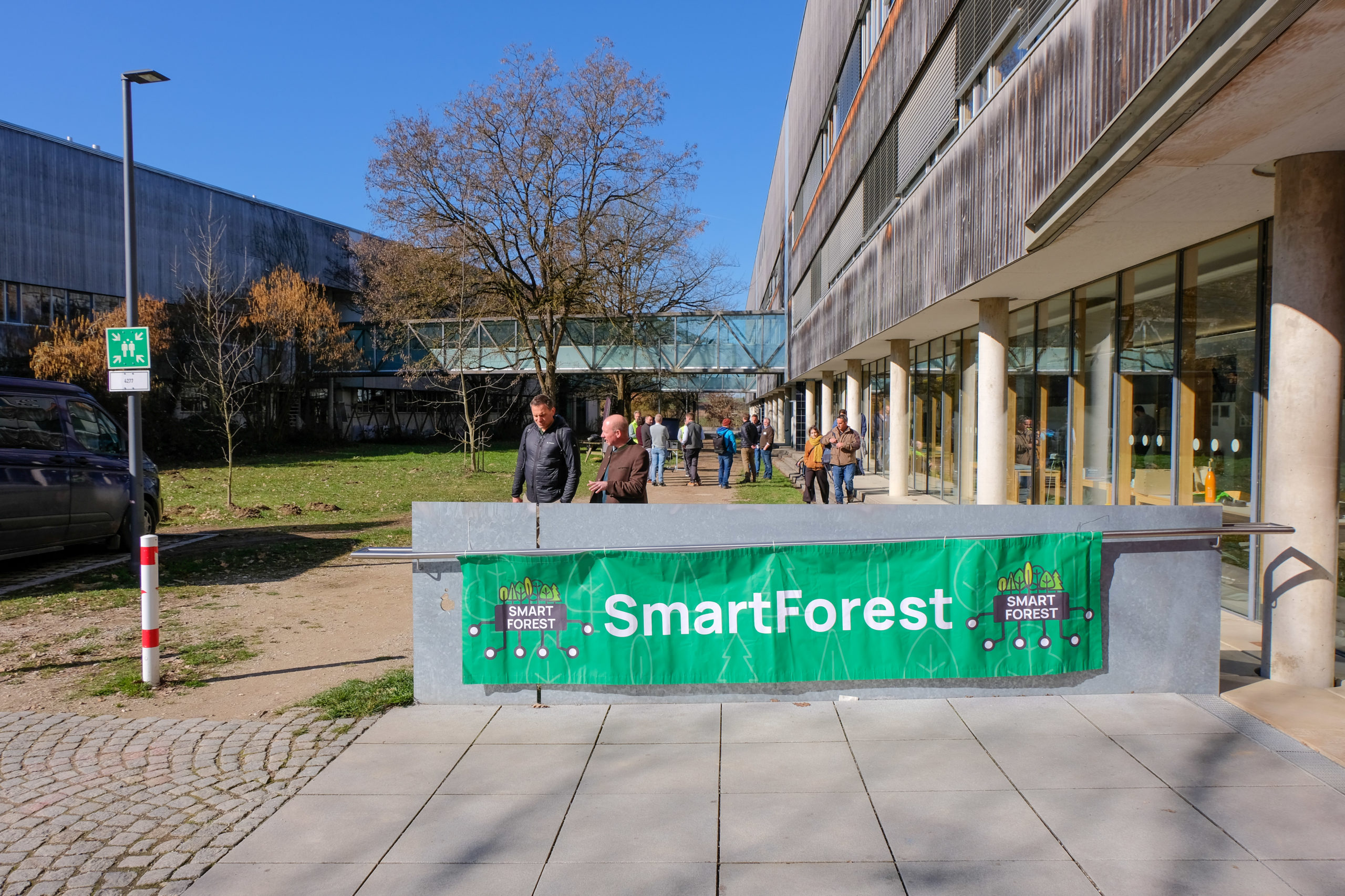 A green, horizontal SmartForest banner hangs outside during the conference. Behind it, various attendees are walking back towards the entrance after a sunny outdoor session in the nearby forest.